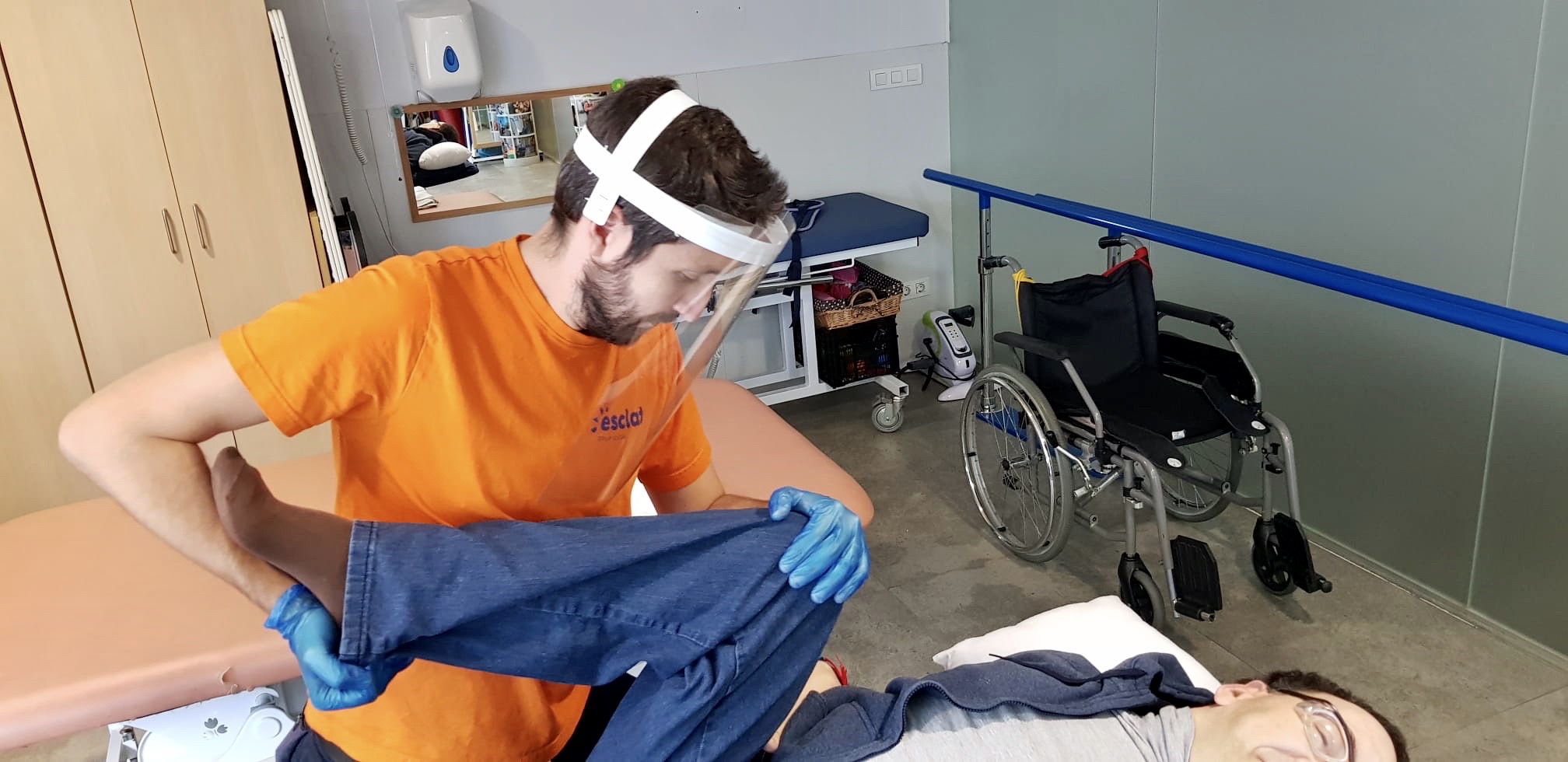 Un fisioterapeuta amb una pantalla de protecció acompanya una persona amb paràlisi cerebral en els seus exercicis terapèutics