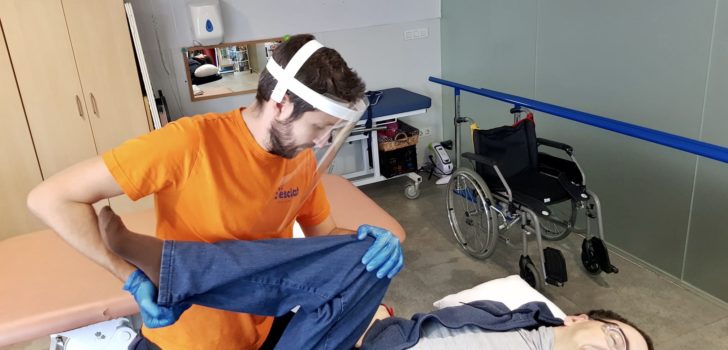 Un fisioterapeuta amb una pantalla de protecció acompanya una persona amb paràlisi cerebral en els seus exercicis terapèutics